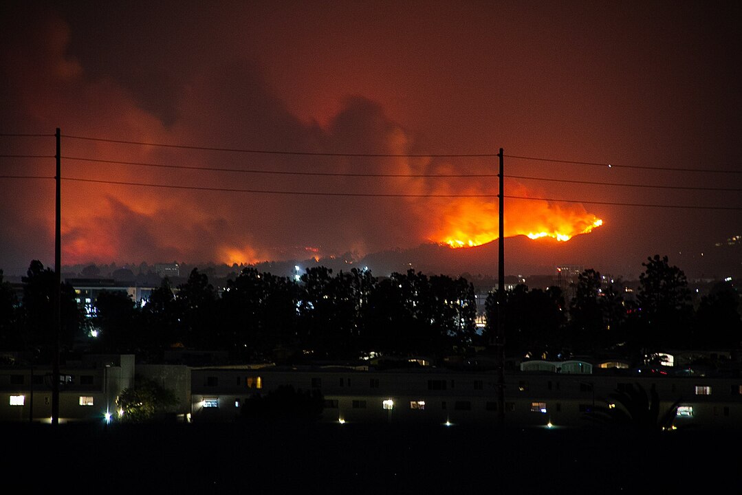 Wildfire from the Palisades Fire as seen from the Playa Vista neighborhood in West Los Angeles on January 7 illuminates the night sky, with burning hills in the background and a silhouette of trees and buildings in the foreground. Photo by FireEditorHomiee/Wikimedia Commons