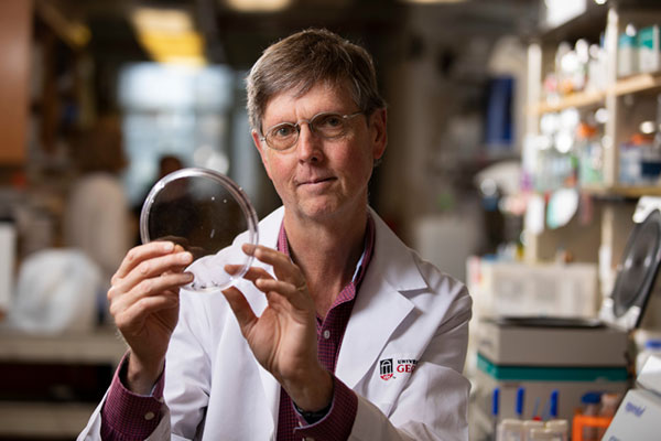 A man with light brown hair and horn-rimmed glasses and a lab coat holds a specimen dish in a lab.