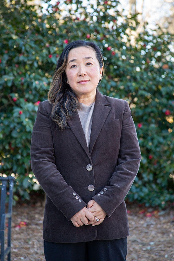 UGA professor Yukiko Hashida stands outdoors in front of leafy greenery with red flowers, wearing a brown blazer and neutral-toned shirt. She has long hair, a calm expression, and clasped hands.