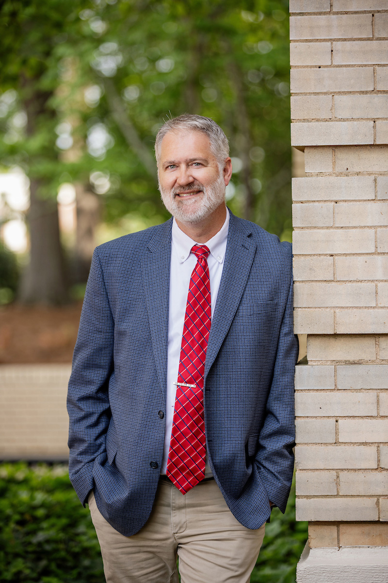 A bearded man leans against a tan brick wall, smiles at the camera.