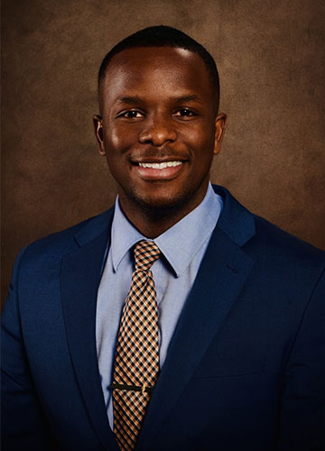 A formal headshot of a smiling young man in a blue sport coat, light blue oxford shirt and blue and yellow patterned tie. 