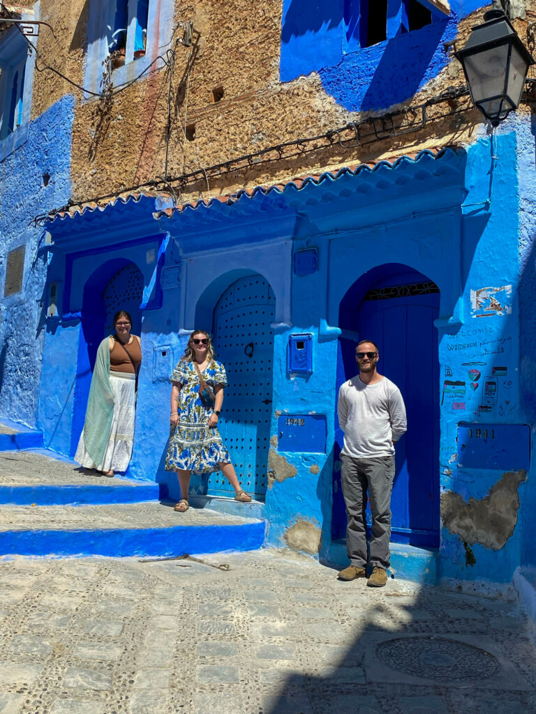Three people pose in front of a sunlit, vibrant blue Moroccan-style building with arched doorways and weathered walls in an alley.