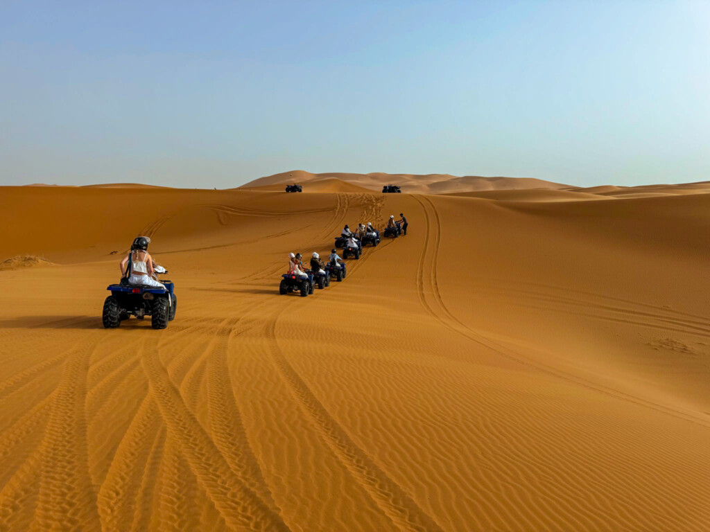 A group of people ride ATVs in a line across a vast sandy desert landscape.