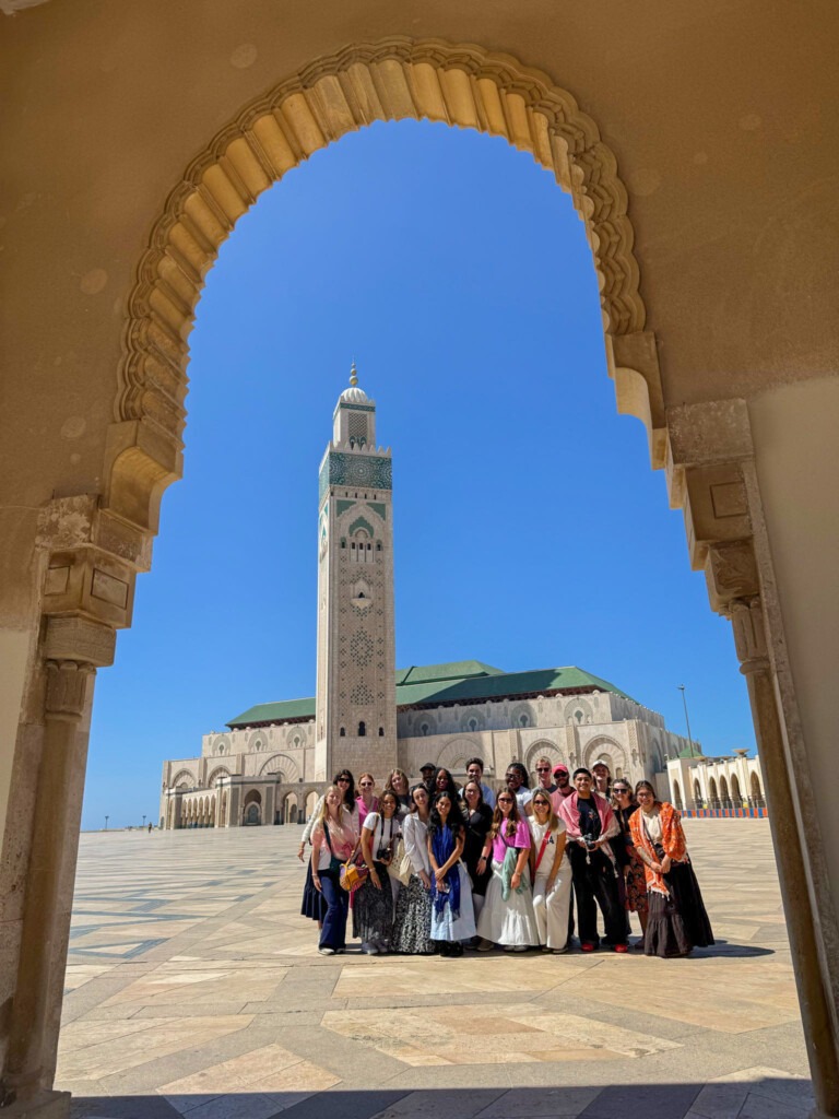 A group of people poses in front of a large mosque with a tall minaret, framed by an ornate arch under a clear blue sky.