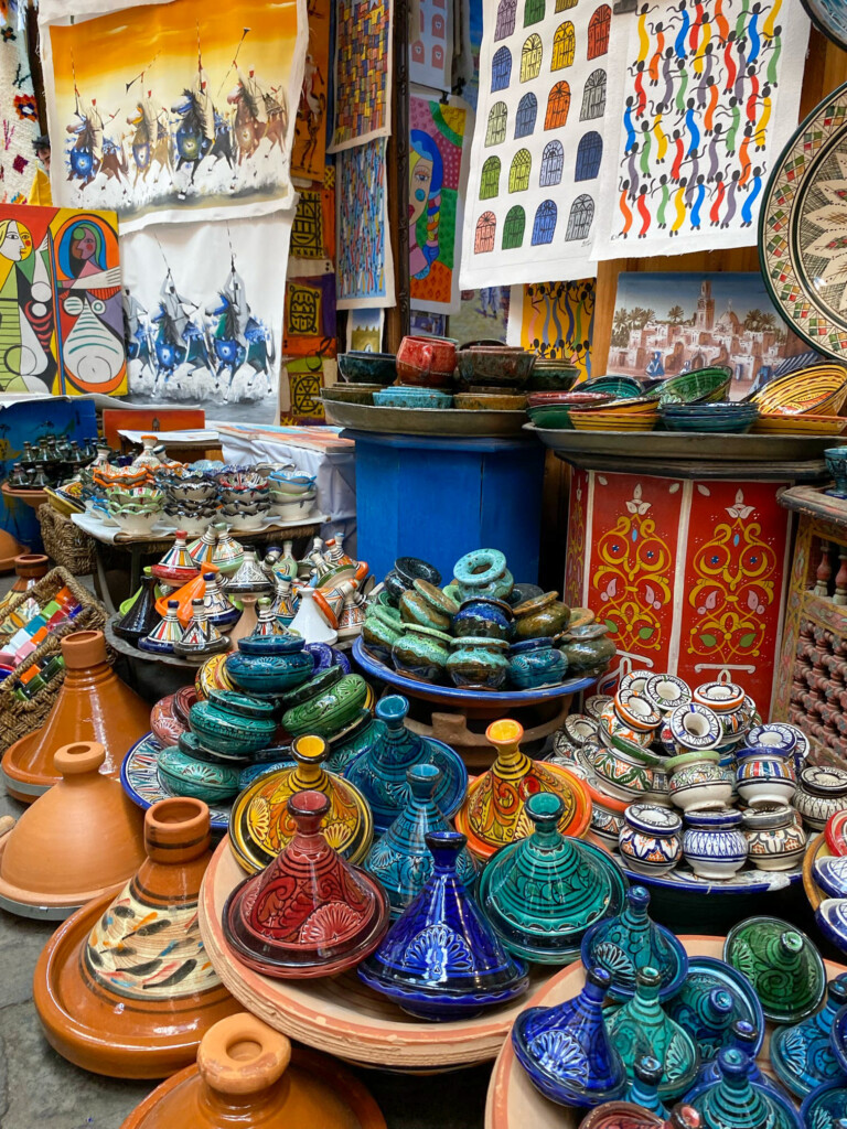 A colorful market stall displays colorful ceramic pottery, including tagines and bowls, alongside patterned textiles and artwork.