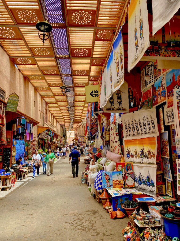 A colorful covered market with stalls selling crafts, textiles, and artwork. Shoppers walk along a stone path under a wooden ceiling with decorative patterns.