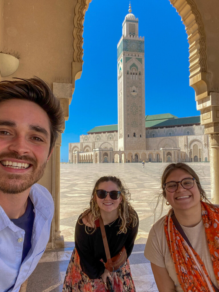 Three people smile for a photo in front of a grand mosque with a tall minaret, framed by an ornate arch under a clear blue sky.