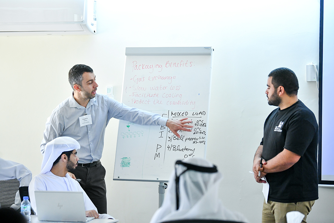 Two men lead a workshop discussion on packaging benefits, presenting to attendees seated near a whiteboard with handwritten notes.