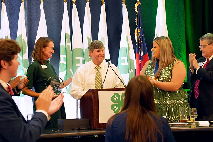 A smiling woman in a green dress presents an award to a smiling mn with salt and pepper hair, a white dress shirt and a green and yellow striped tie at a formal Georgia 4-H event. 