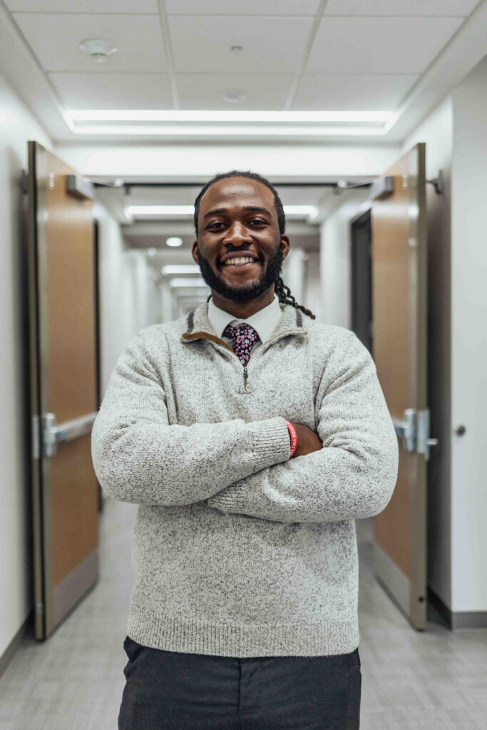 A Black male poses for a photo in a hallway, smiling with his arms crossed.