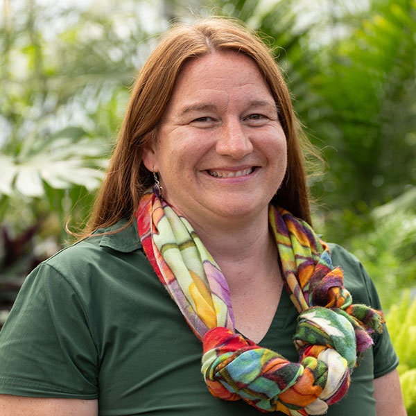 Julie Campbell, wearing a green shirt and colorful scarf, smiles in front of palms in a greenhouse