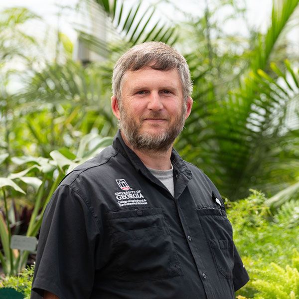Ben Campbell, wearing a shirt branded for the UGA College of Agricultural and Environmental Sciences, smiles in front of palms in a greenhouse