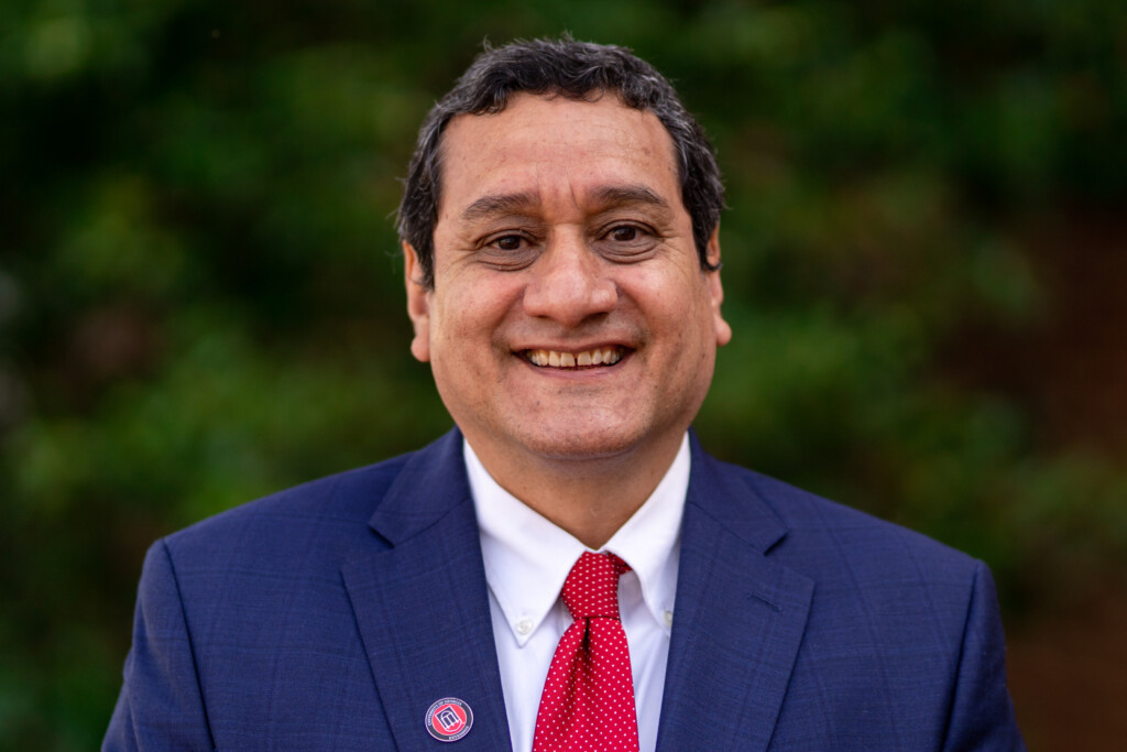 Alfredo Martinez, wearing a suit and tie, smiles in front of greenery outdoors