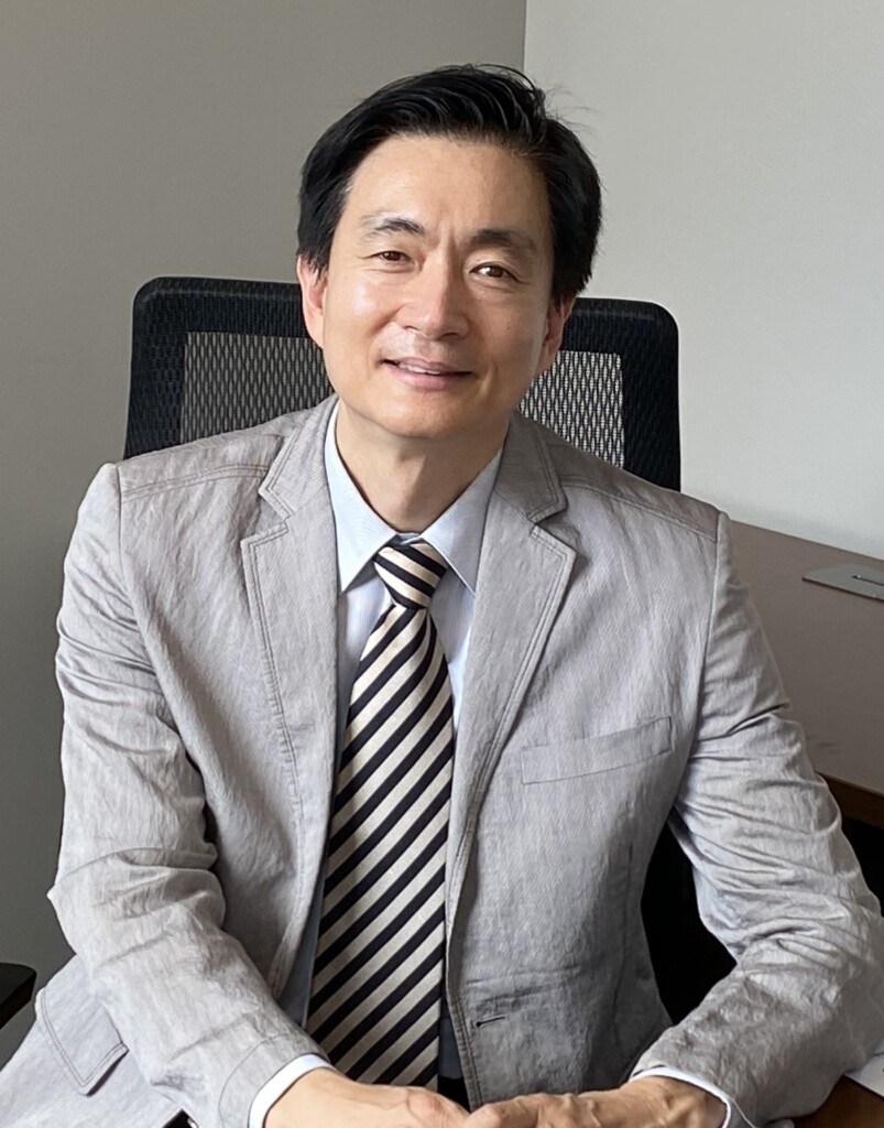 Woo Kyun Kim, wearing a suit and tie, smiles in an office setting