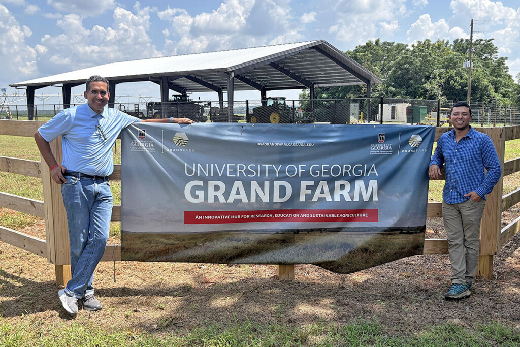 Two men stand on either side of a blue banner with white lettering hung on a fence in front of a shelter holding farm equipment. The sign reads University of Georgia Grand Farm, An innovative hub for research, education and sustainable agriculture.
