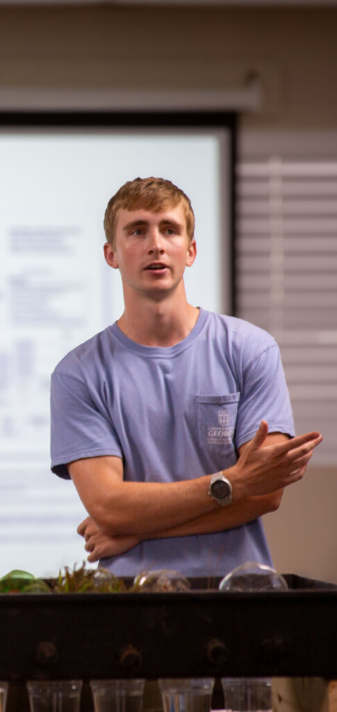 A young man stands at the front of a classroom, arms loosely crossed, speaking to the group. A projected slide behind him reads “Soil Tests from UGA” with bullet points about plant nutrients. In front of him sits a demonstration table filled with soil samples, clear cups, and teaching materials. Bright indoor lighting highlights his pale blue T-shirt and the informal, hands-on learning setting.