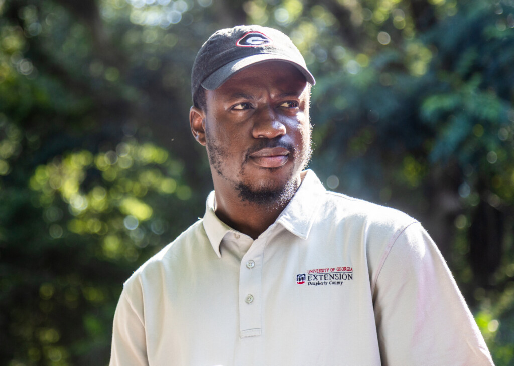 A man wearing a University of Georgia Extension polo and a black UGA cap stands outdoors in bright, dappled sunlight. He looks slightly to the side with a focused expression, with blurred green foliage creating a soft backdrop behind him.
