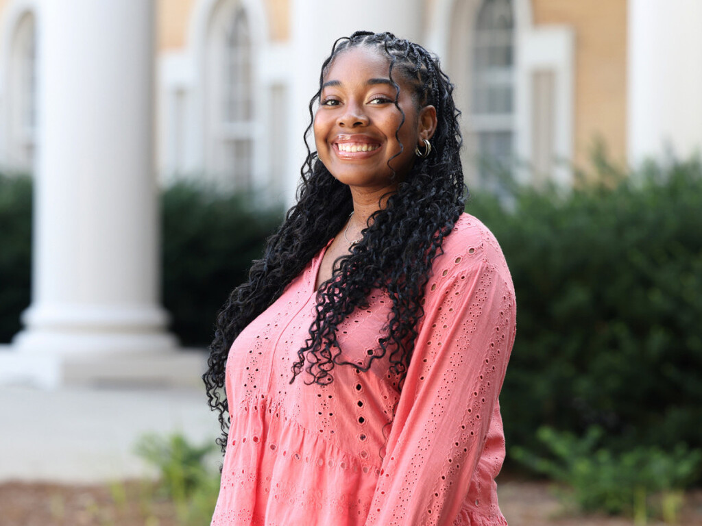 A young woman with long, curly braids smiles while standing outdoors in front of tall white columns and greenery. She wears a coral pink, eyelet-patterned blouse.