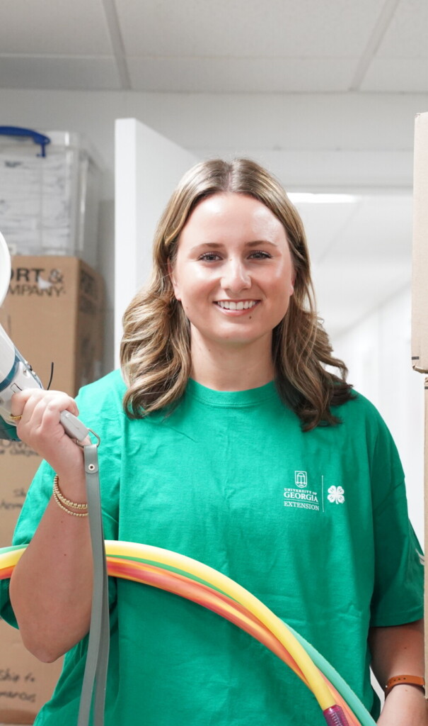 A young woman wearing a green UGA Extension 4-H T-shirt smiles while holding a megaphone and several colorful hula hoops. She stands in a storage area lined with cardboard boxes, including large Amazon boxes stacked to one side.