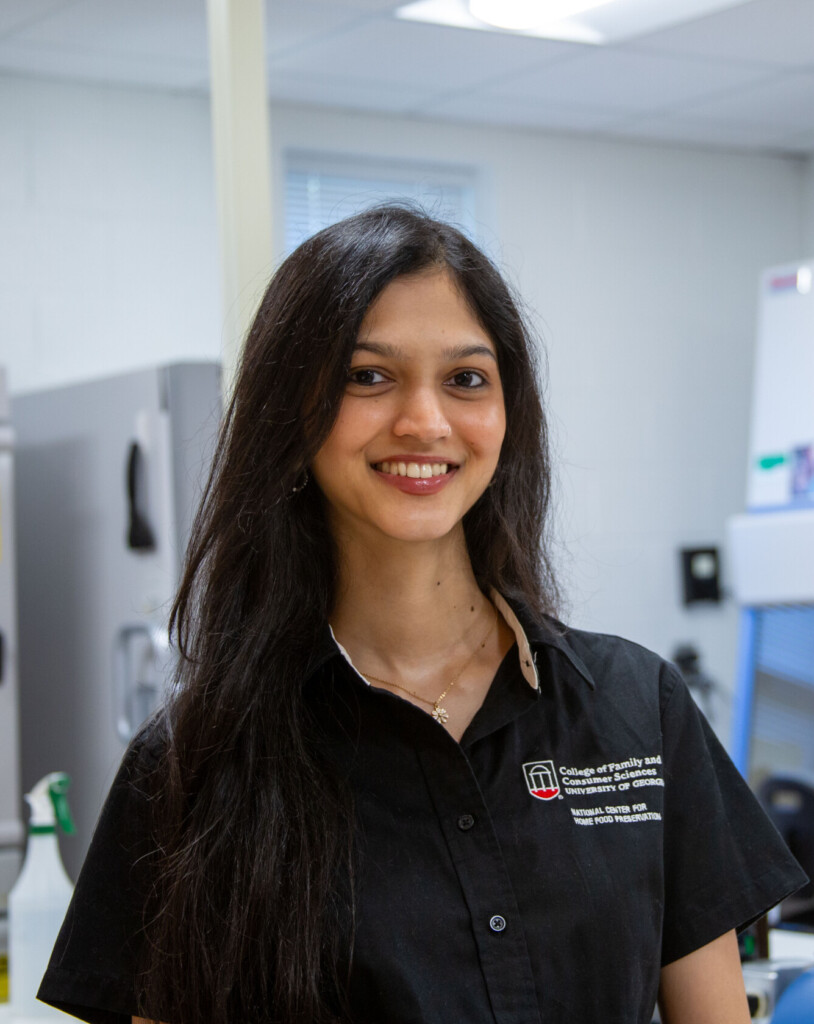 A woman wearing a black University of Georgia College of Family and Consumer Sciences shirt smiles while standing in a laboratory. Behind her are lab equipment, including incubators and a biosafety cabinet, along with various instruments on the counter.