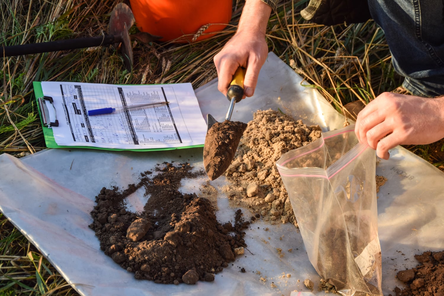 a person shovels soil from two different piles into a plastic bag while filling out a soil testing form