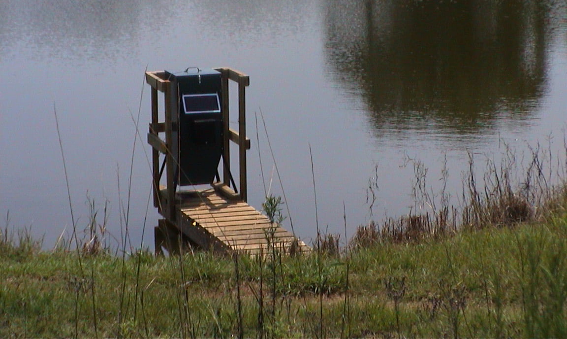 A fish feeder placed on a dock for easy access and activated by a timer and solar power.