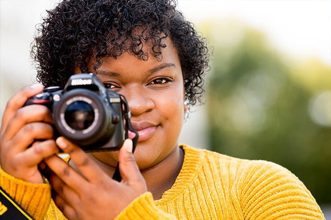 a Black woman in a yellow sweater holds a camera up close to her face