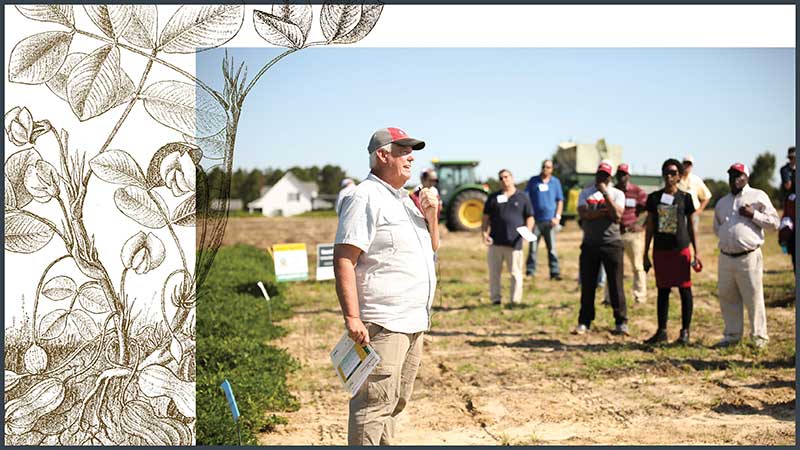 A man in a ball cap, blue shirt and tan pants stands in a field talking to a group of listeners.