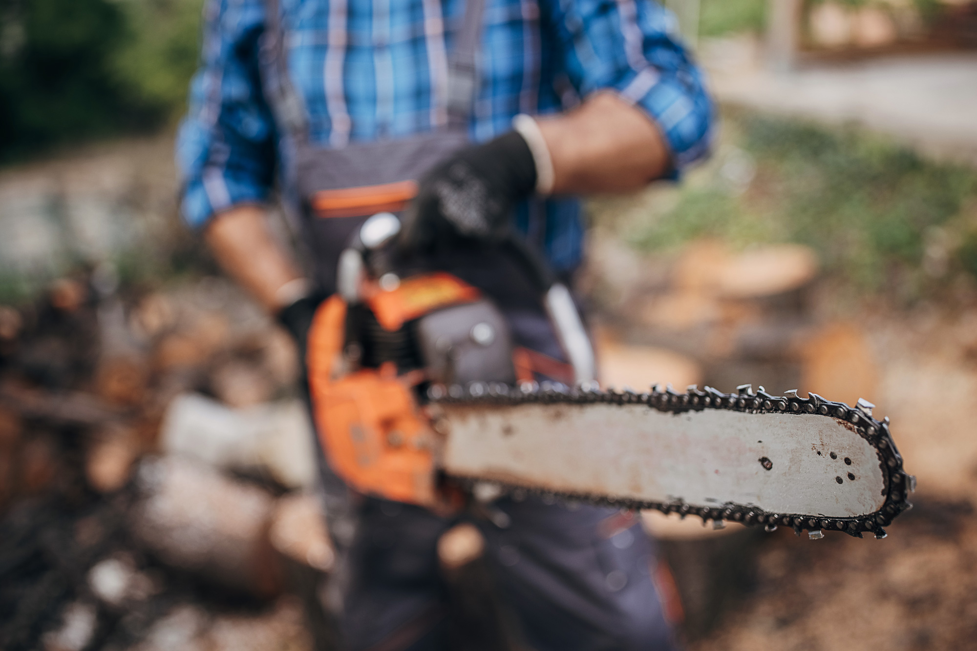 A man stands in the background wearing gloves and holding a chainsaw toward the camera, with the focus on the chainsaw's chain