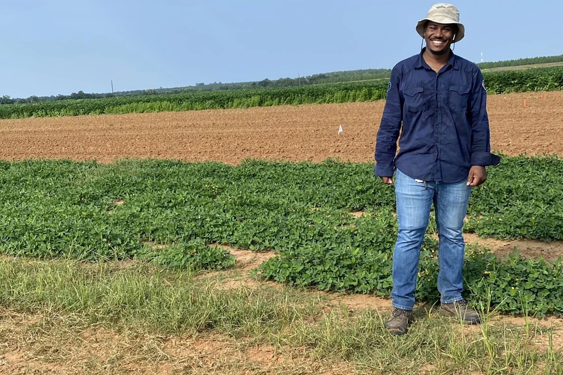 A man in a sun hat, navy button up shirt and jeans stands in a field, smiling for the camera
