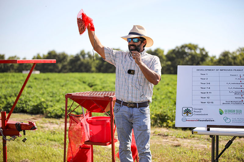 A man in a sun hat and sunglasses, wearing a short-sleeved plaid shirt and jeans, holds up a red net bag holding peanuts while standing in front of a peanut field surrounded by equipment.