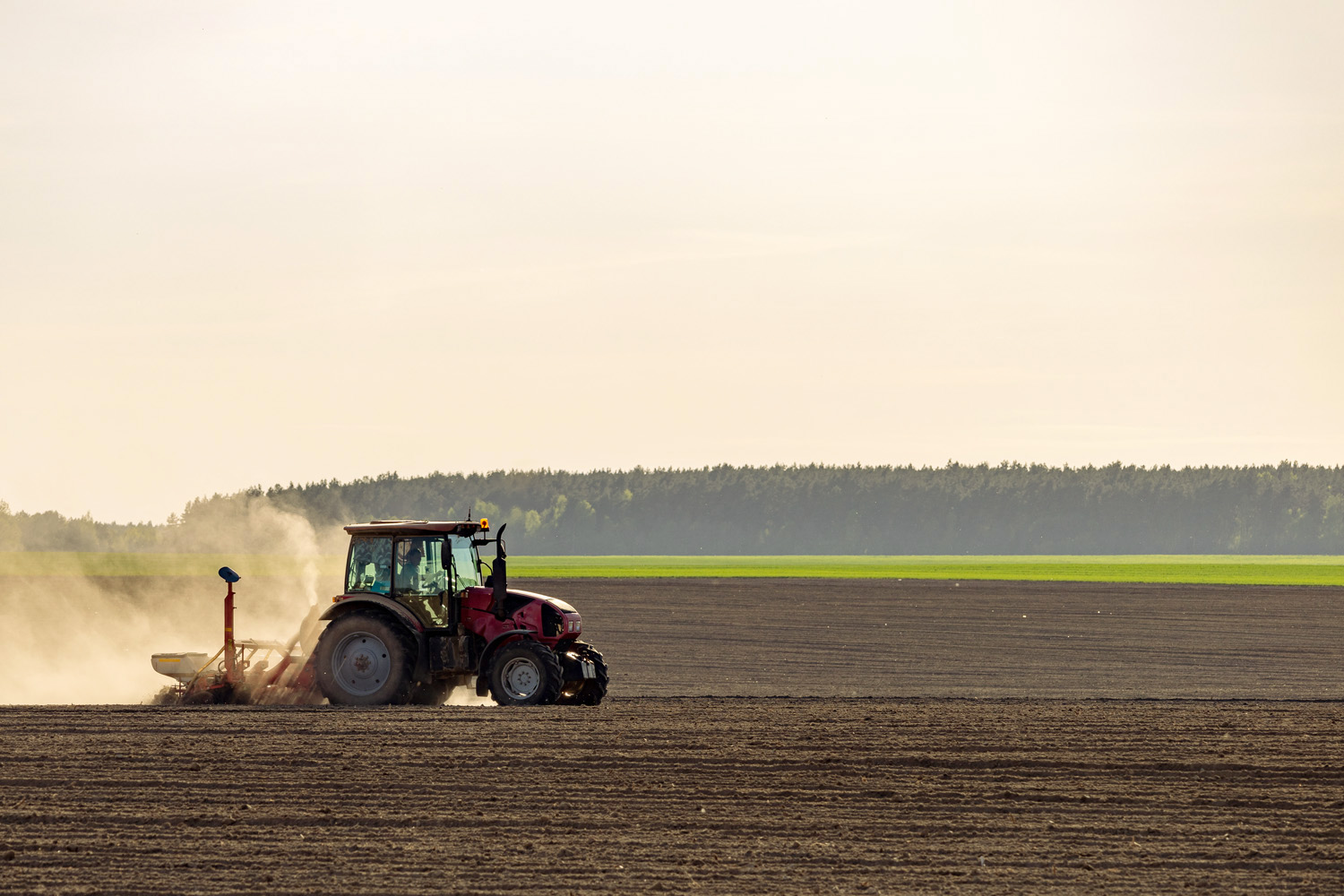 A tractor works in a ploughed field with green fields and trees in the distance