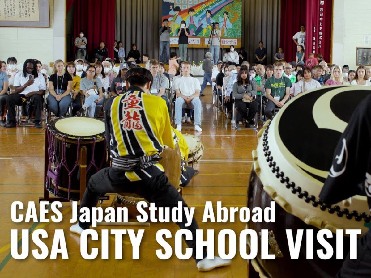 An assembled crowd watching a performance in a school auditorium. The words "CAES Japan Study Abroad: Usa City school visit" are overlayed in white.