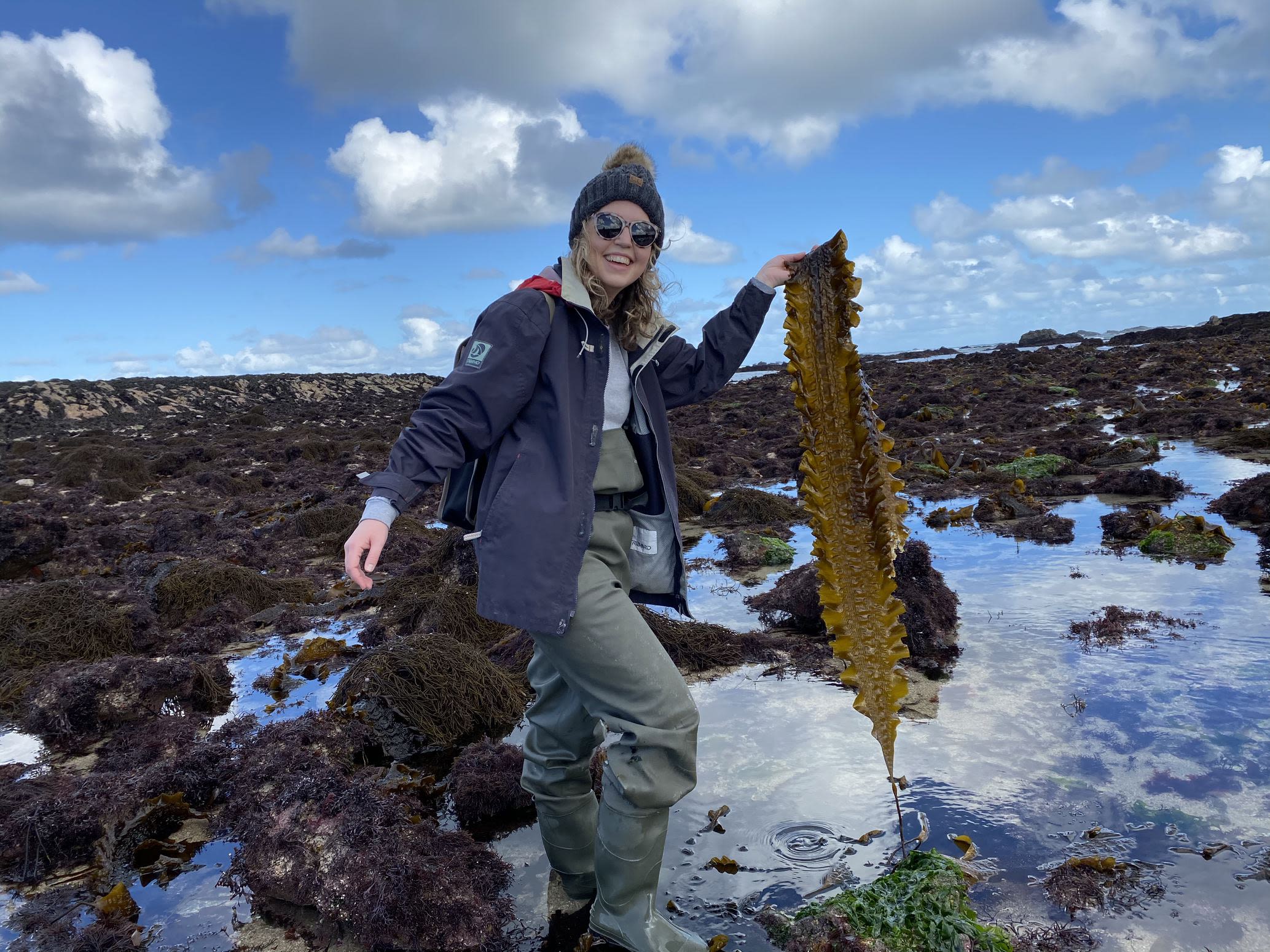 A woman stands in a field of wild seaweed holding a large piece of seaweed and smiling for the camera.