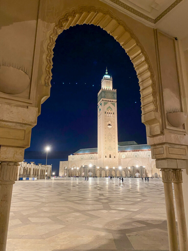 A grand mosque with a tall illuminated minaret is framed by an ornate arch at night, with a spacious patterned courtyard and people walking below.