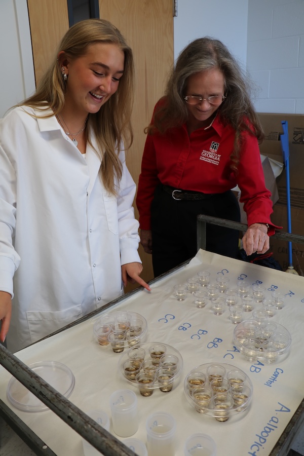 Sydnie Frost (left) stands in a white lab coat and Nancy Hinkle (right) stands in a red button up and black pants. The two look over a cart with small clear plastic cups and labels and smile while they discuss the experiment.