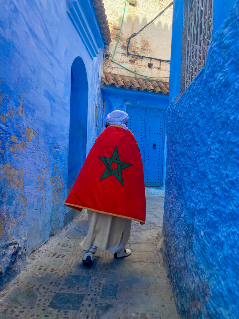 Person wearing a Moroccan flag cape walks down a narrow blue alleyway.