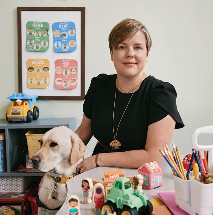 A woman in a black shirt sits next to a yellow Labrador. In front of them is a table covered with children's toys. Behind them is a all with more toys and children's educational posters.