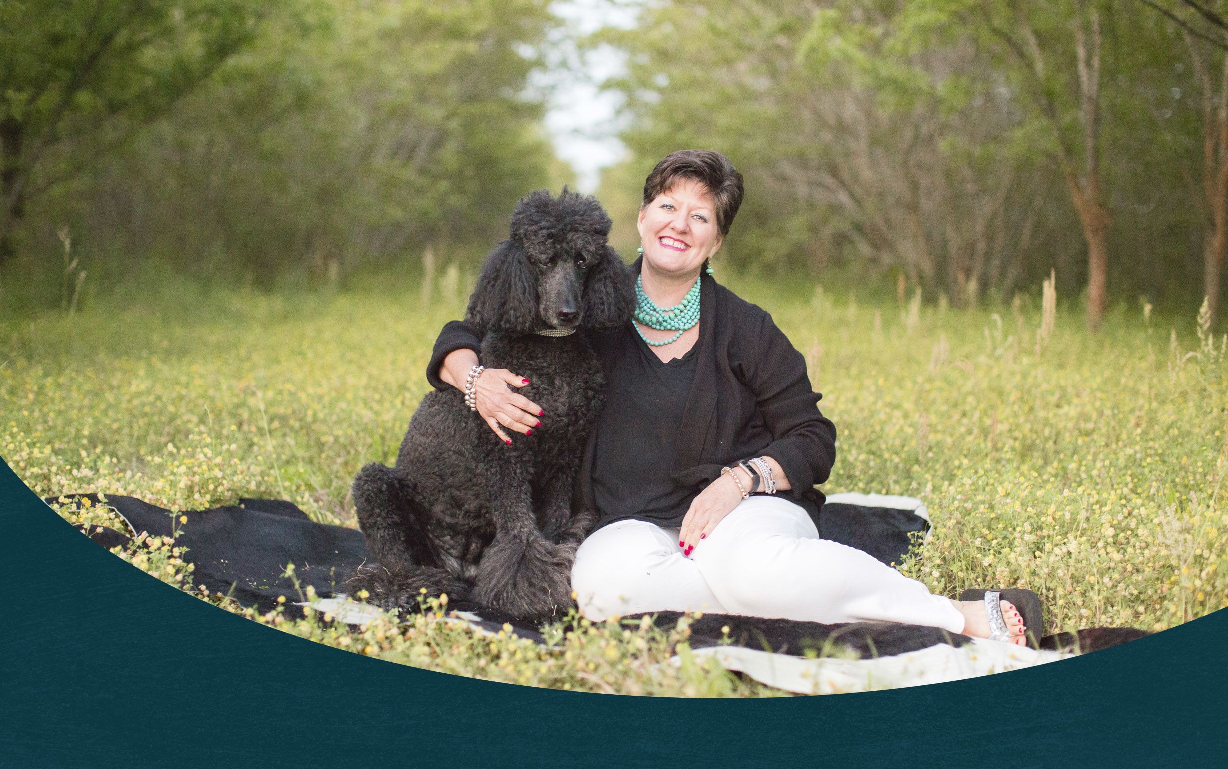 Laura Perry Johnson smiles while she sits in an orchard with her arm around her black poodle. Johnson wears an aquamarine beaded necklace and a black long-sleeve shirt.