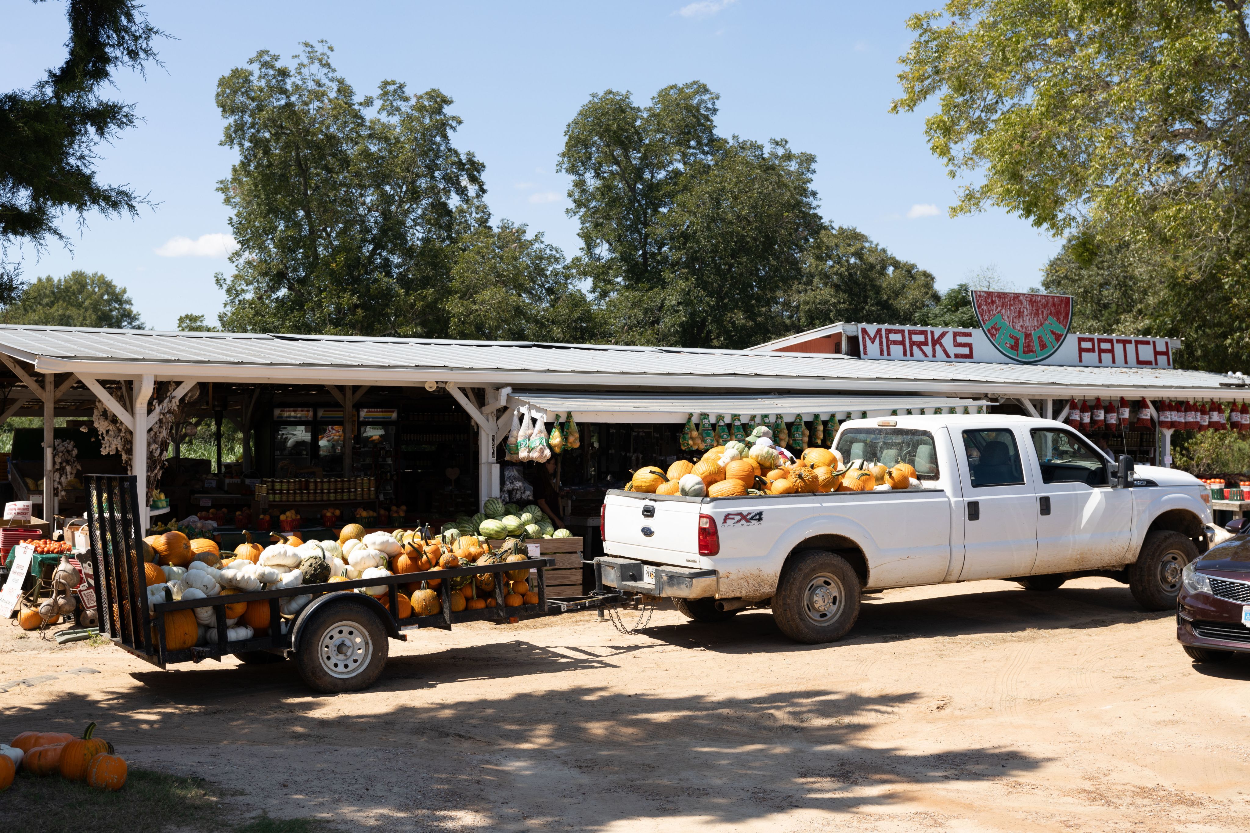 A white truck full of pumpkins pulls a trailer full of pumpkins is parked in front of the farm stand. 