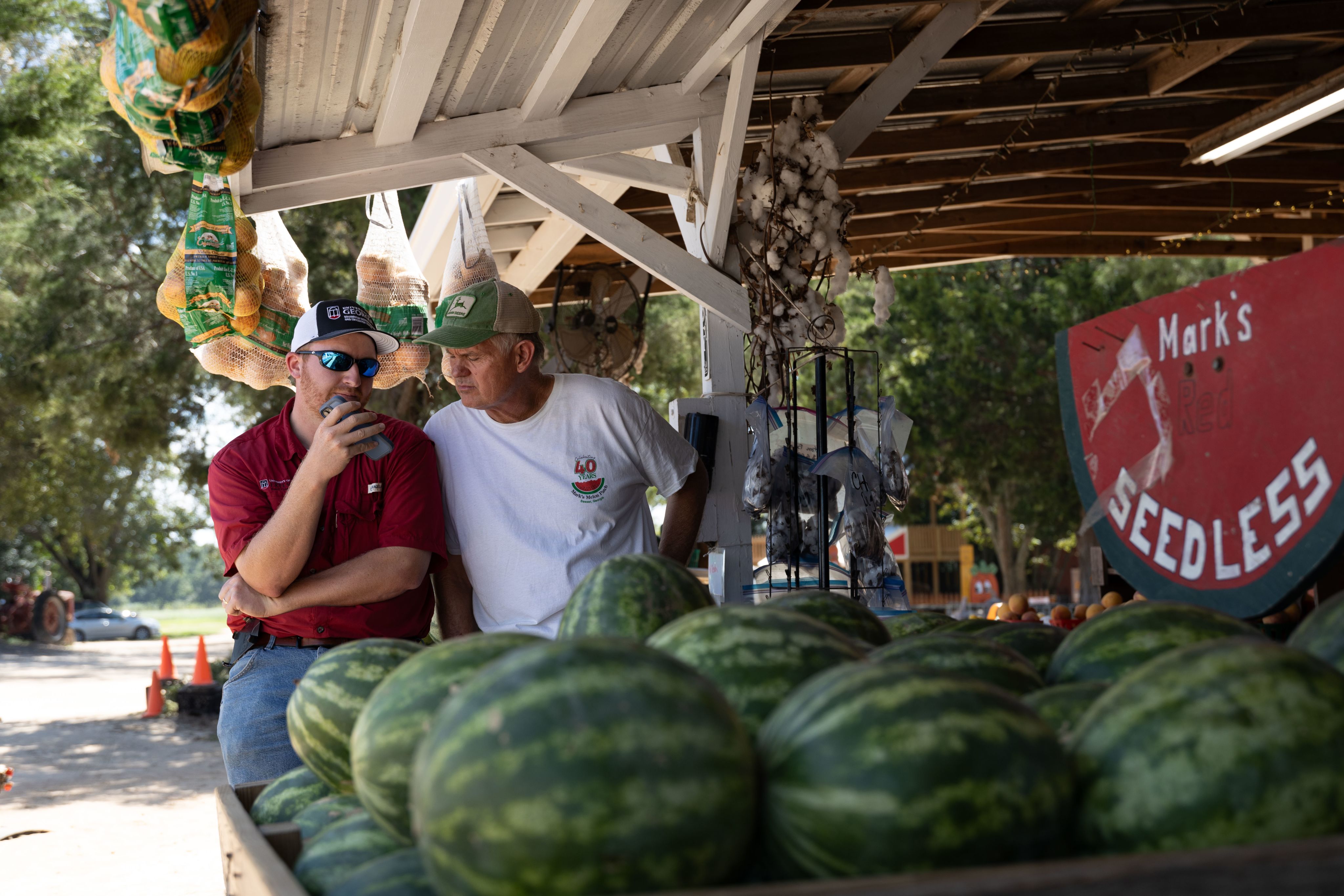 McAllister on the left in a red shirt, hat and sunglasses sits next to Mark Daniel, who's wearing a white shirt and green and beige hat. McAllister holds his phone while the two talk on the speaker phone. Watermelons can be seen in the foreground and bags of onions hang from the ceiling of the farm stand.