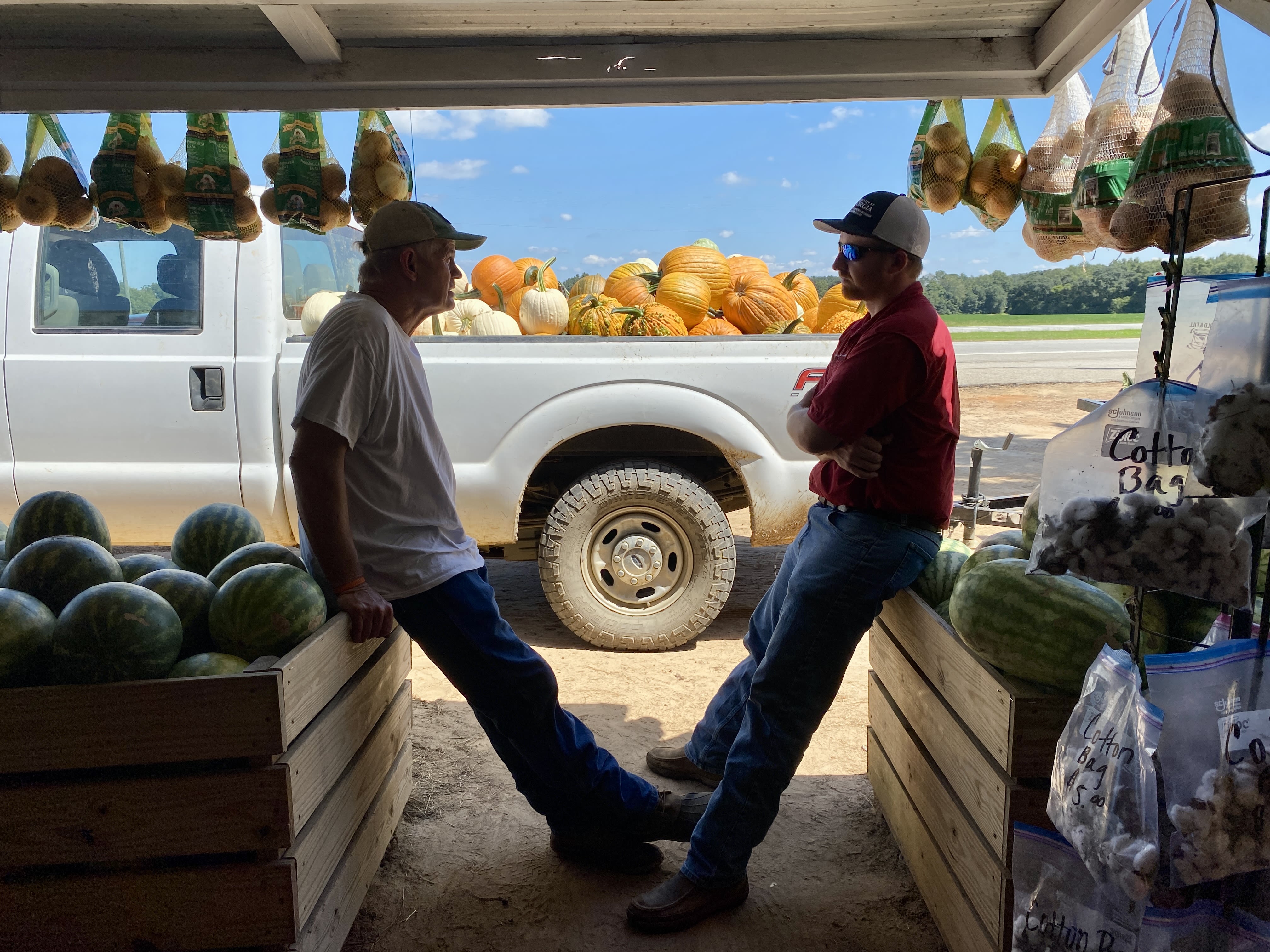 Mark Daniel on the left leans against a wooden bin full of watermelons while looking at Seth McAllister who is also leaning against a wooden bin of watermelons. A white truck in the background is full of orange pumpkins. 