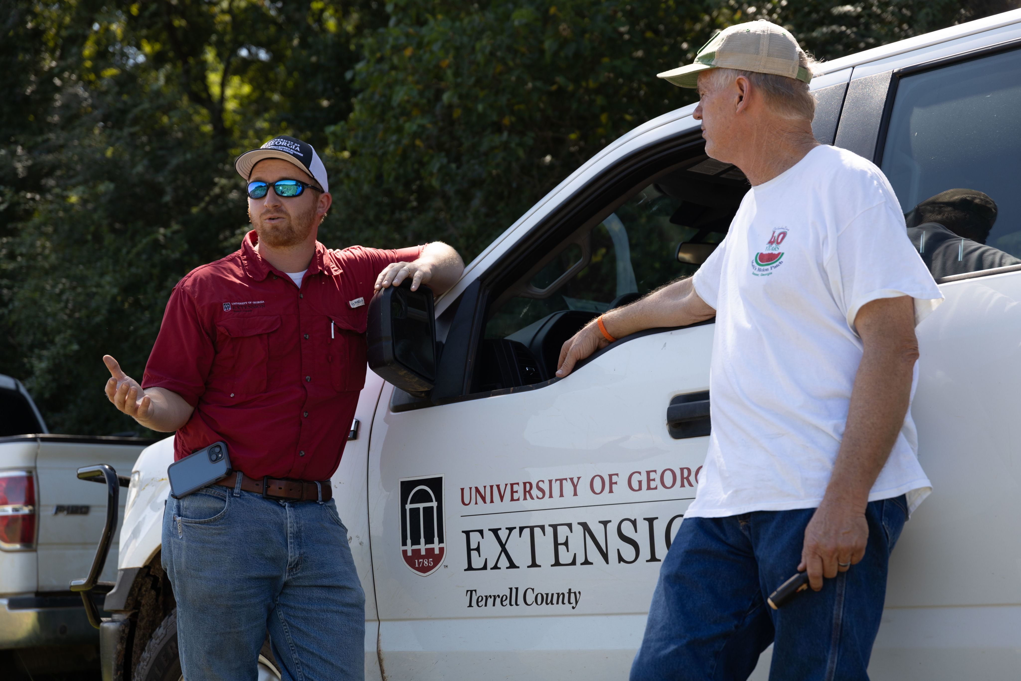 Seth McAllister on the left wears a red shirt, sunglasses and a hat while he's standing in front of a white truck while talking to Mark Daniel who is wearing a beige hat and white shirt.