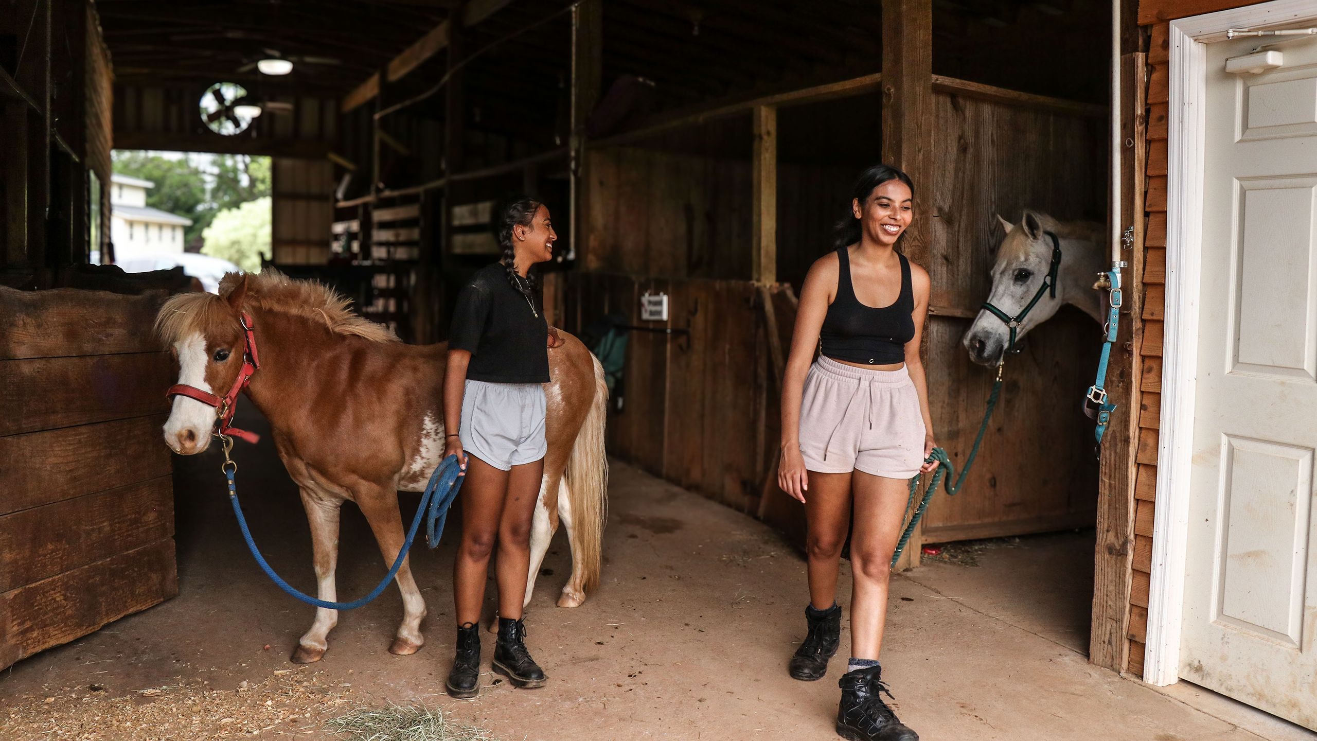 Two girls lead horses out of a stable with lead rope