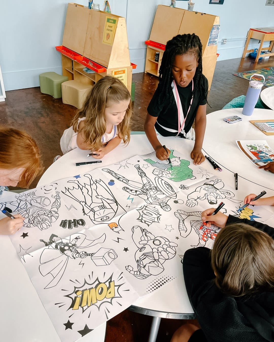 Several children stand at a table in a classroom setting and color coloring pages.