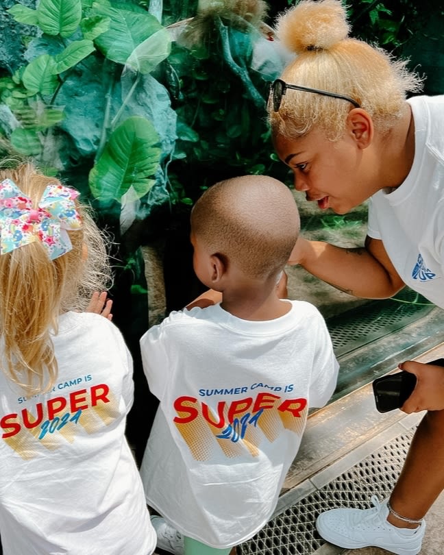 A woman points towards plants with two children looking on