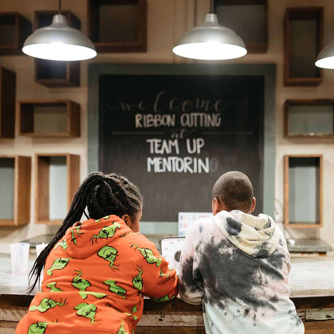Two students sit at a desk behind a chalkboard with "Welcome Ribbon Cutting at Team Up Mentoring" written on it.