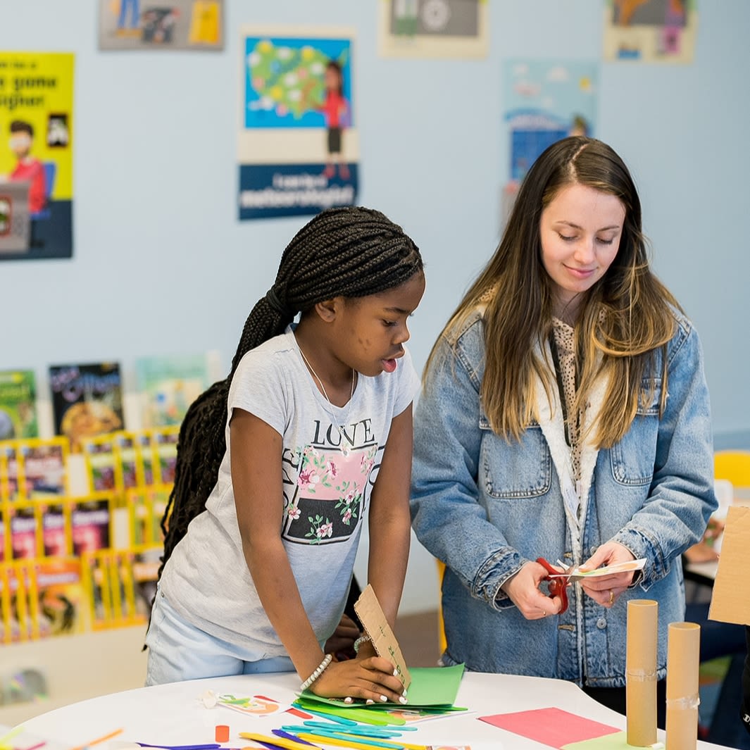 Two young women stand in a classroom setting looking down at craft supplies. One cuts a piece of paper with craft scissors.