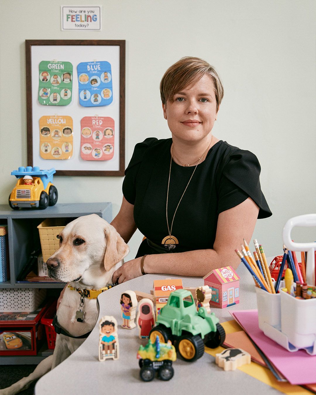 A woman in a black shirt sits next to a yellow Labrador. In front of them is a table covered with children's toys. Behind them is a all with more toys and children's educational posters.