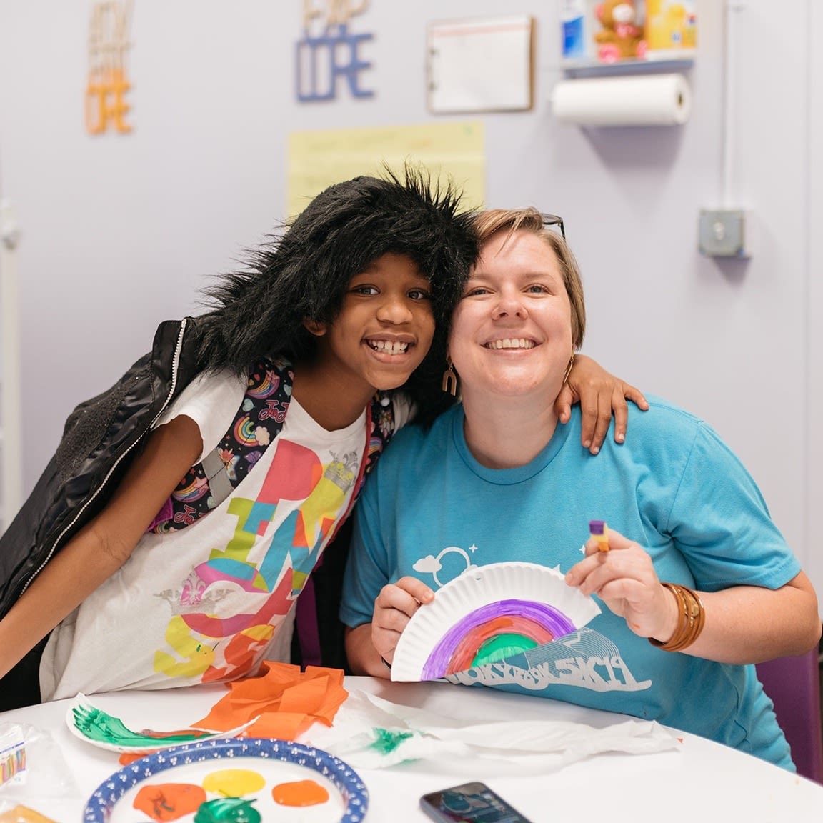 A young girl smiles at the camera with her arm around a woman to her right. The woman holds up a painted paper plate and sits behind a table covered in craft supplies.